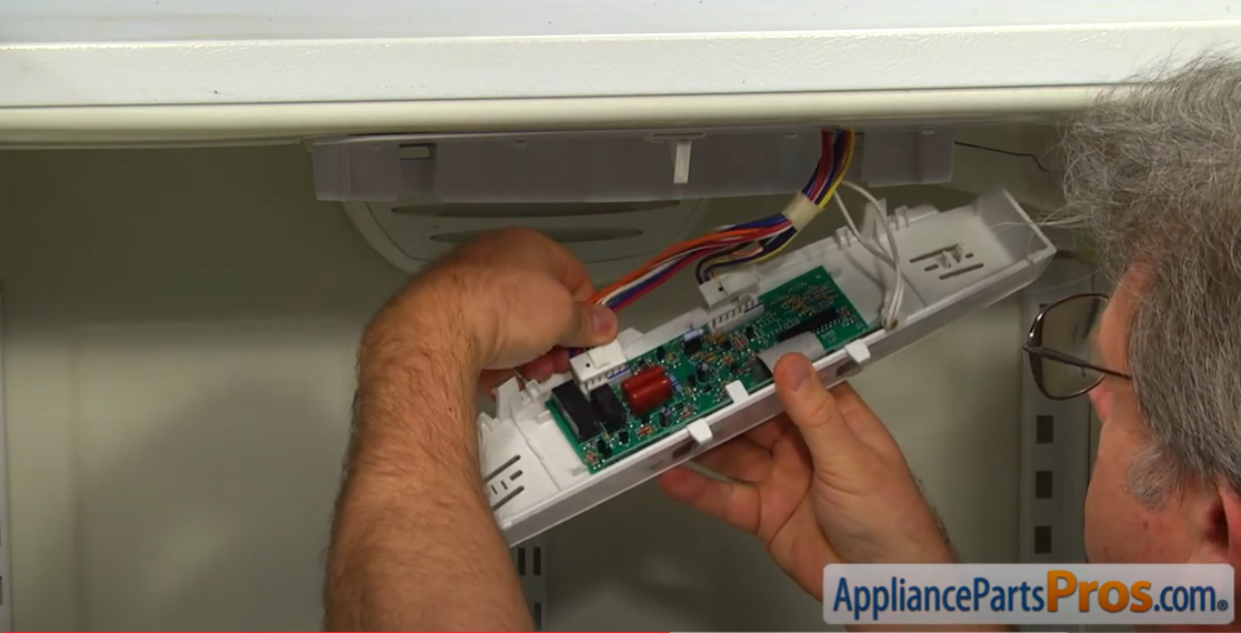 Hands connecting a wire harness to a new temperature control board in a fridge housing