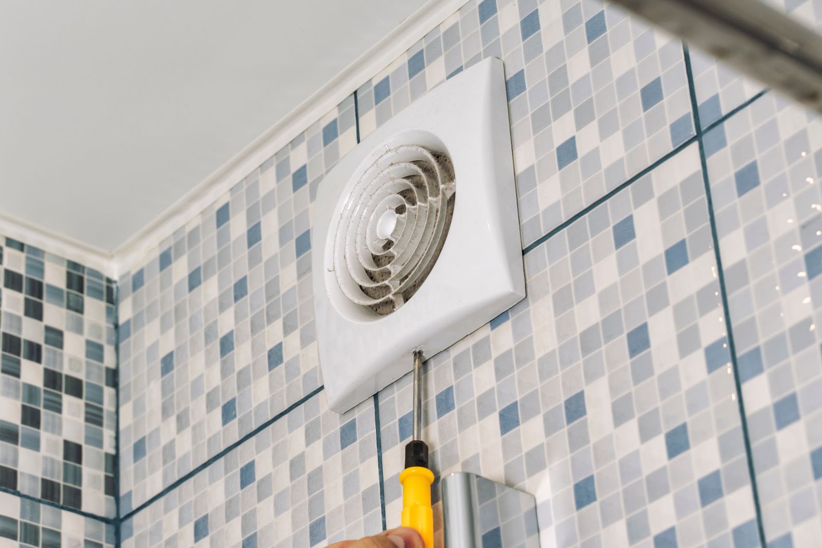 Screwdriver at the base of a white bathroom exhaust fan cover on a tiled wall
