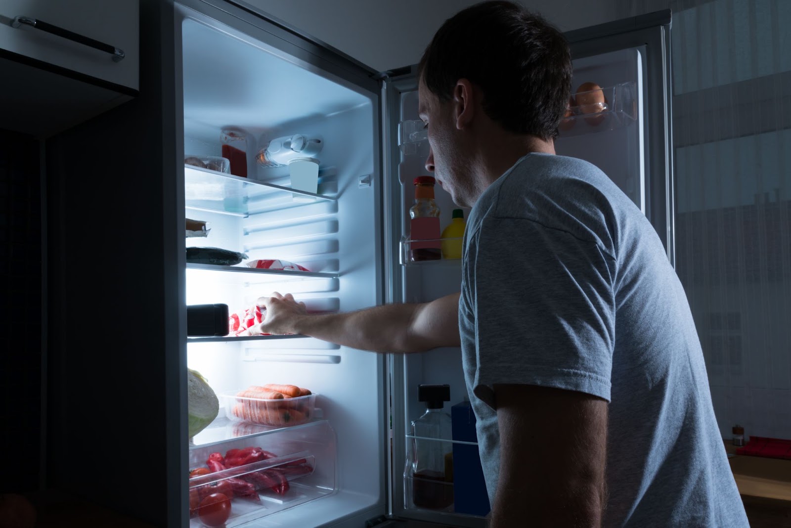 Man reaching into a lit refrigerator in a dark kitchen