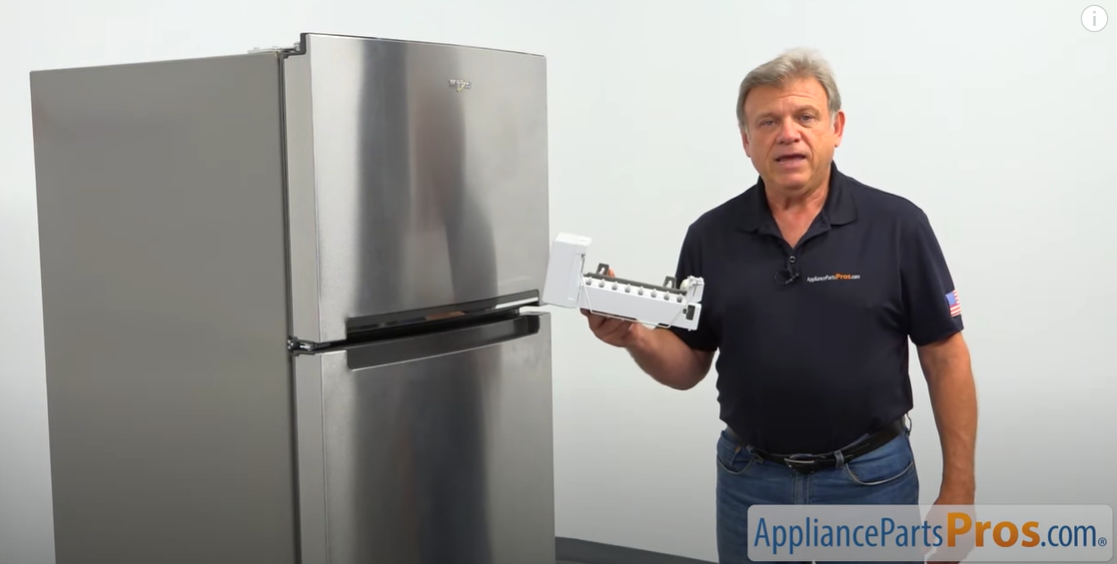 Technician holding an ice maker assembly beside a stainless top-freezer refrigerator
