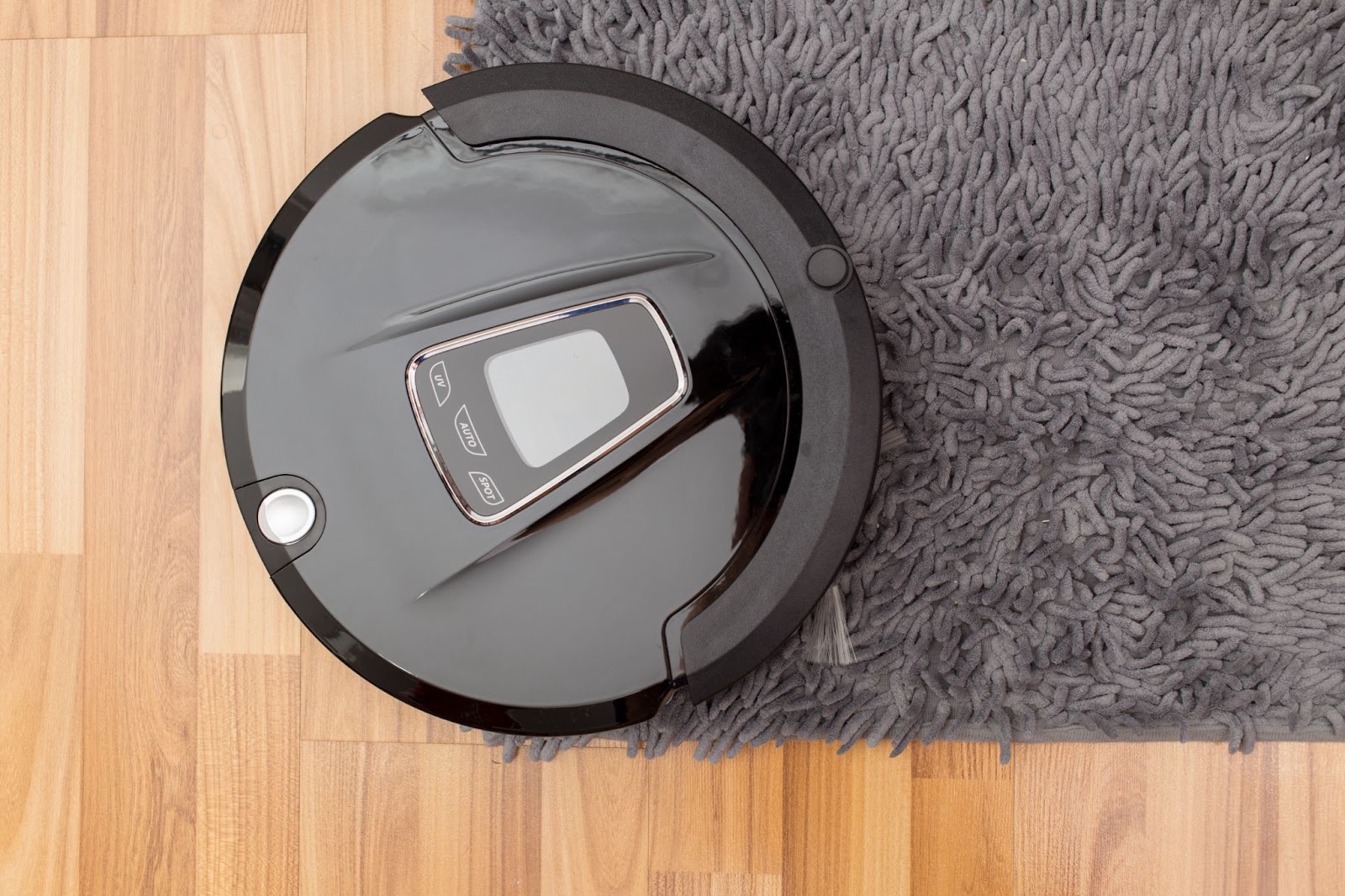 Top view of a round black robot cleaner on a wood floor at the edge of a gray rug